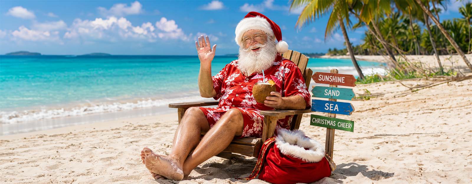 Image of Santa Claus waving while sitting on a beach wearing shorts.
